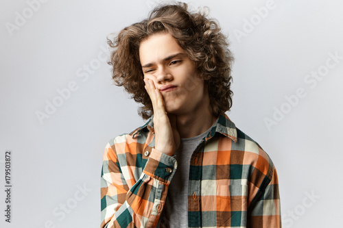 People, pain and sickness concept. Studio shot of upset frustrated young male with painful look, having terrible toothache because of cavity, holding hand on his cheek, doesn't want to go to dentist