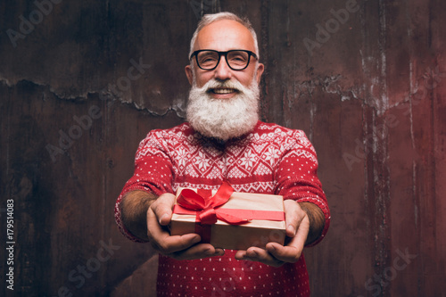 Senior bearded man holding gift box over dark background and looking at camera with smile  Santa Claus wishes Merry Christmas and a happy new year 2018!