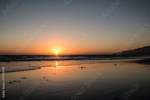 Fototapeta Naklejka Na Ścianę i Meble -  Red Yellow Orange Sunset Over Water With Waves On Shoreline Wet Sand On Beach Reflecting Sunset