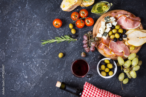 Fotografia Appetizer, italian antipasto, ham, olives, cheese, bread, grapes, pear and wine on dark stone background