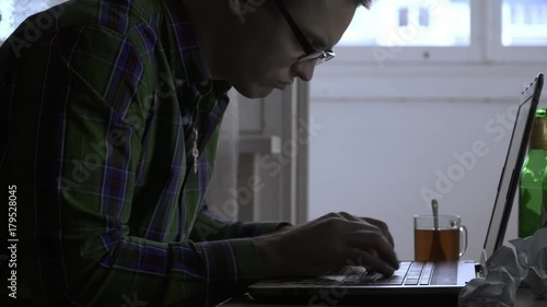 A writer or a programmer works at home for a laptop not for the first day. A man gets angry and starts smoking a cigarette. On the table are bottles with beer, tea, cigarettes, paper photo