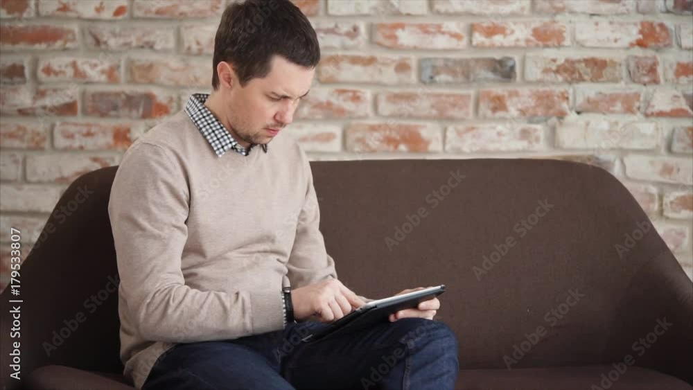An adult man who sits on the couch is copied on the tablet with a business partner, persons holding a gadget in his hand and printing text An adult man who sits on the couch is copied on the tablet with a business partner, persons holding a gadget in his hand and printing text