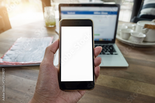 Close up of a man holding smartphone with blank screen mobile and laptop computer at coffee shop.