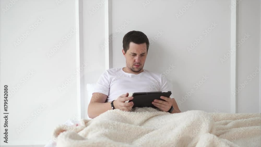 Man is lying in big bed under white duvet with a tablet after a long day. He is surfing in the internet, reading news and watching video. Man is lying in big bed under white duvet with a tablet after a long day. He is surfing in the internet, reading news and watching video.
