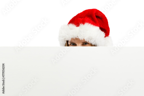 Beautiful woman in Santa Claus clothes peeking behind blank board on white background