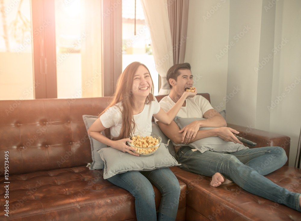 Young couple relaxing in their apartment, lying on the couch, watching a movie and eating popcorn. Lens flare effect on the window, Young couple relaxing in their apartment, lying on the couch, watching a movie and eating popcorn. Lens flare effect on the window,