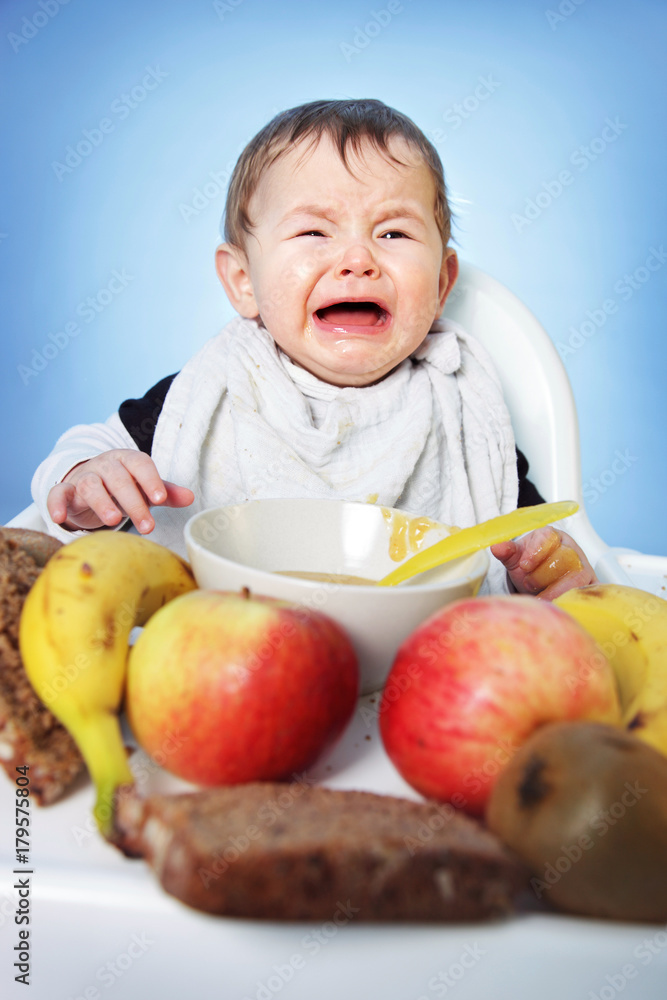 Crying unhappy Baby boy must eat healthy food for breakfast on bright blue background Crying unhappy Baby boy must eat healthy food for breakfast on bright blue background