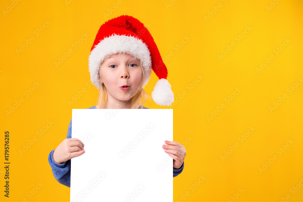 Cute little girl in Santa Claus hat with a white blank paper sheet. Christmas, x-mas, New Year, Winter, people, advertisement, sale concept. Cute little girl in Santa Claus hat with a white blank paper sheet. Christmas, x-mas, New Year, Winter, people, advertisement, sale concept.
