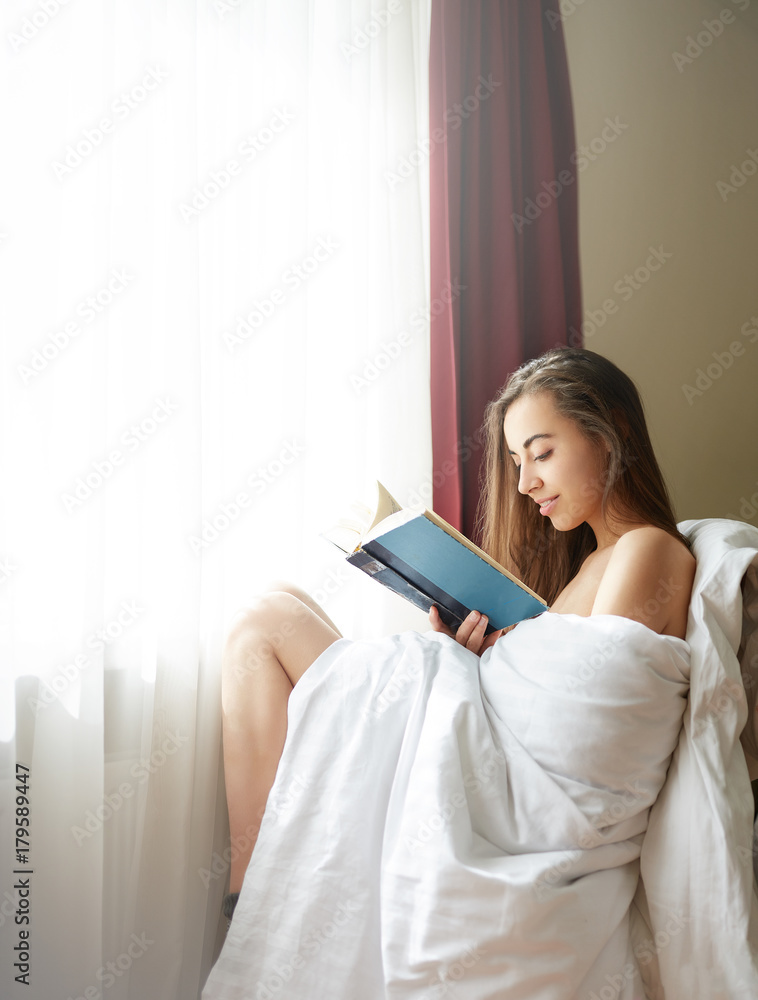 young smiling woman sits near a window with old book. the woman is sitting without clothes, in warm socks and wrapped in a blanket young smiling woman sits near a window with old book. the woman is sitting without clothes, in warm socks and wrapped in a blanket