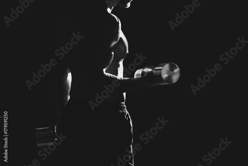 Silhouette of a slender bodybuilder with heavy dumbbells in his hands, doing sports in the gym.