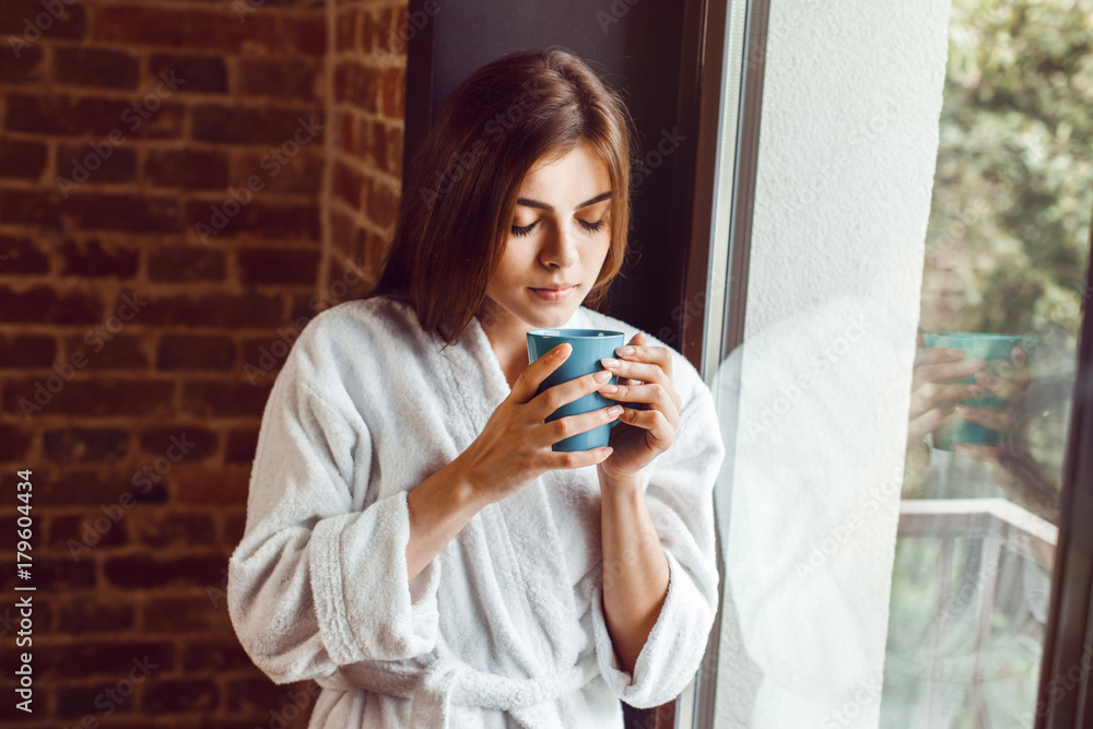 Beautiful caucasian girl, in a white bathrobe, enjoys morning coffee time in the kitchen near window Beautiful caucasian girl, in a white bathrobe, enjoys morning coffee time in the kitchen near window