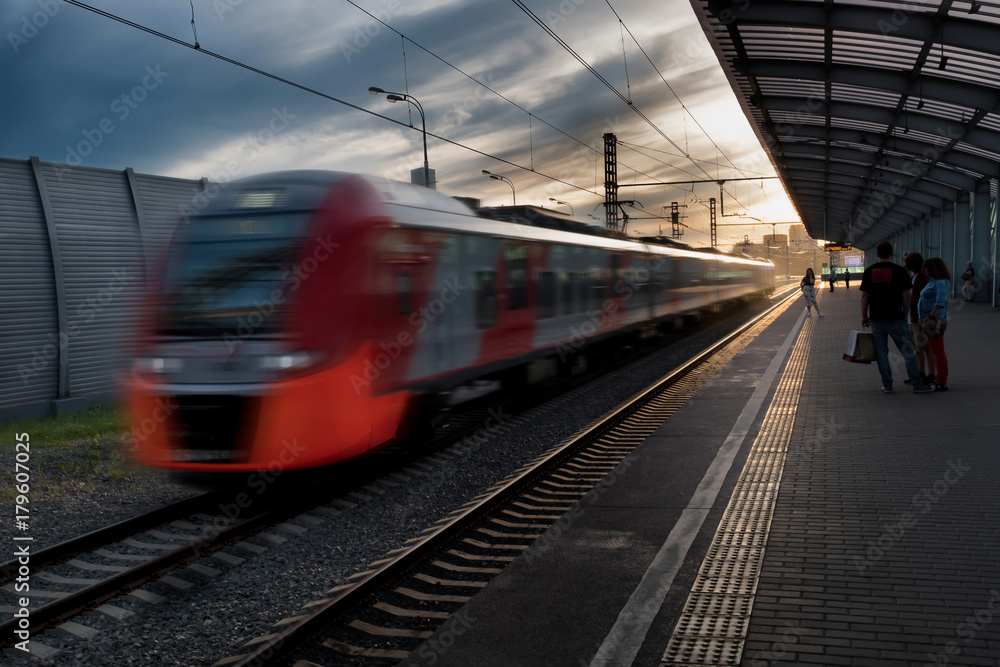 Naklejka premium High-speed beautiful red modern train in motion at the railway station at sunset in Europe. Blur effect. Industrial scene with a passenger train on the railway on the sunset