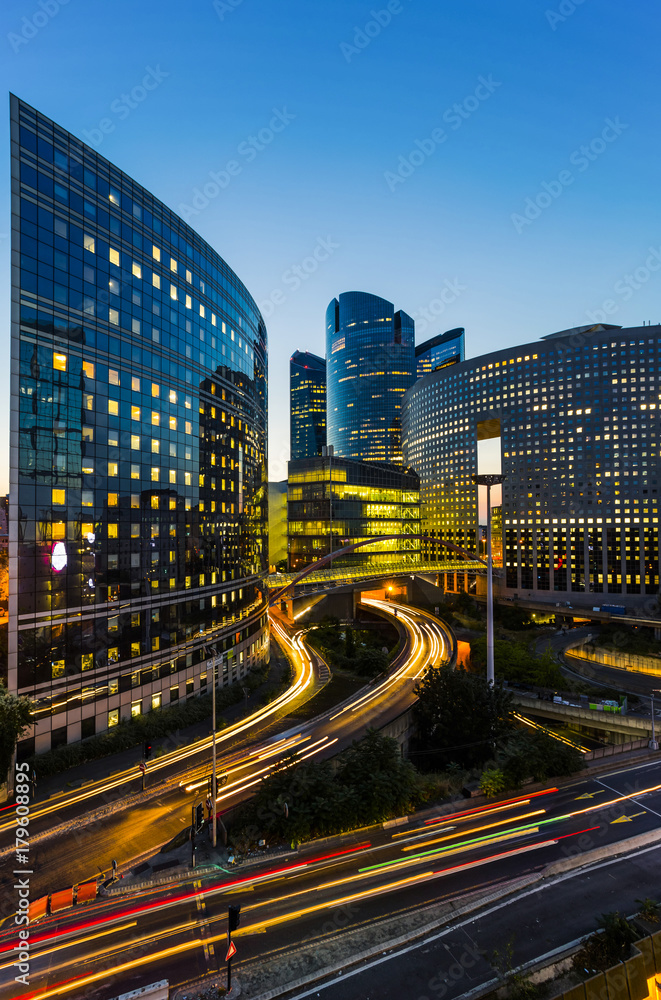 Fototapeta premium Skyscrapers in Paris business district La Defense. European night cityscape with dynamic street traffic, car lights and glass facades of modern buildings. Economy, finances, transport concept. Toned