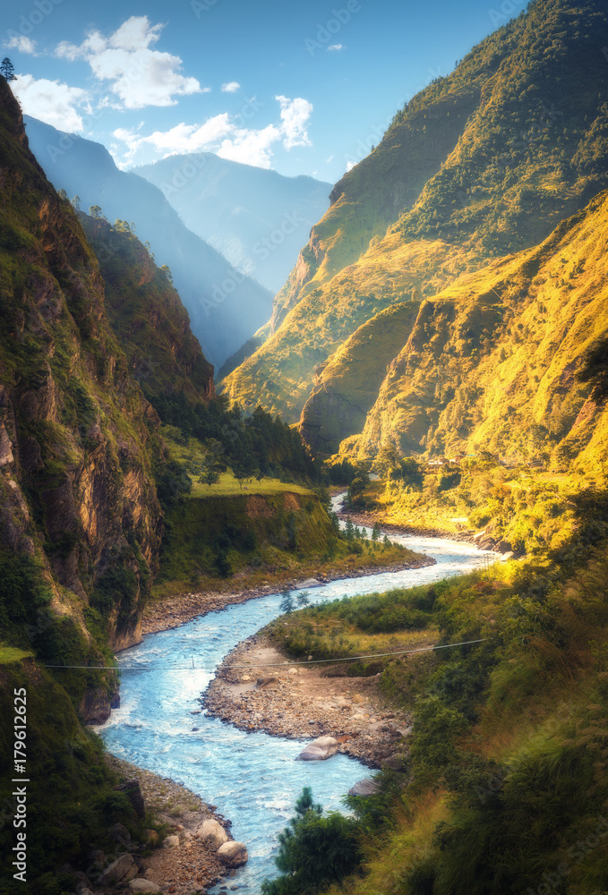 Naklejka premium Niesamowity krajobraz z wysokimi Himalajami, piękną krętą rzeką, zielonym lasem, błękitnym niebem z chmurami i żółtym słońcem jesienią w Nepalu. Górska dolina. Podróżuj po Himalajach. Natura