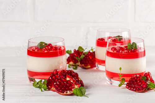 Creamy vanilla panna cotta with red jelly in beautiful glasses, fresh ripe pomegranate on white wooden background.