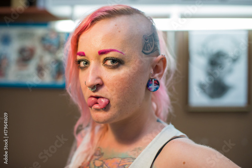
A young girl with a shaved head, pink hair and a tattoo on his head and chest. Many piercings on the face and body. Tunnels in the ears. photo