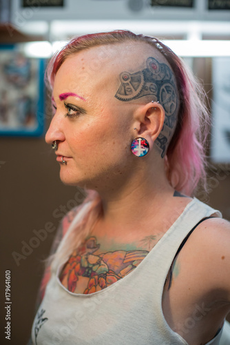 
A young girl with a shaved head, pink hair and a tattoo on his head and chest. Many piercings on the face and body. Tunnels in the ears. photo