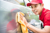 Asian man worker in a red uniform happily cleaning a car with a yellow microfiber cloth