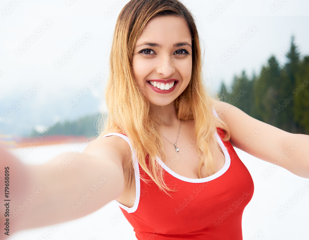 Selfie portrait of young cheerful woman skier wearing red swimsuit, taking a selfie on snowy slope at winter ski resort. People, lifestyle and technology concept Selfie portrait of young cheerful woman skier wearing red swimsuit, taking a selfie on snowy slope at winter ski resort. People, lifestyle and technology concept