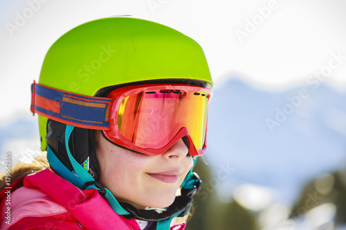 Portrait of happy young girl sitting in the snow with ski in winter time, ski slope in the background.
