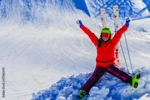 Portrait of happy young girl sitting in the snow with ski in winter time, ski slope in the background.