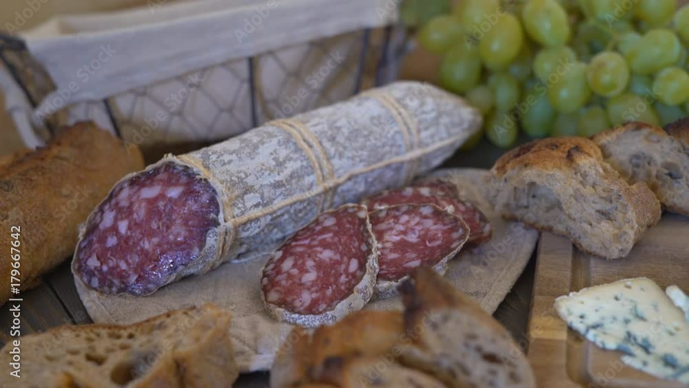 appetizing sausage loaf in a bag of marble fabric. the sausage rests on a wooden table of brown color. In the background a baguette, cheese with green mold and a large bunch of white grapes with a appetizing sausage loaf in a bag of marble fabric. the sausage rests on a wooden table of brown color. In the background a baguette, cheese with green mold and a large bunch of white grapes with a