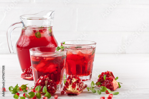 Pomegranate cocktail with ice and mint in beautiful glasses and jug, fresh ripe pomegranate on white wooden background. Sweet red juice. Close up photography. Selective focus