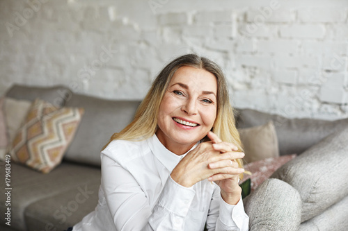 People, age, maturity and happiness concept. Indoor portrait of happy attractive senior woman with healthy skin and loose hair sitting on big grey couch, looking at camera with joyful smile