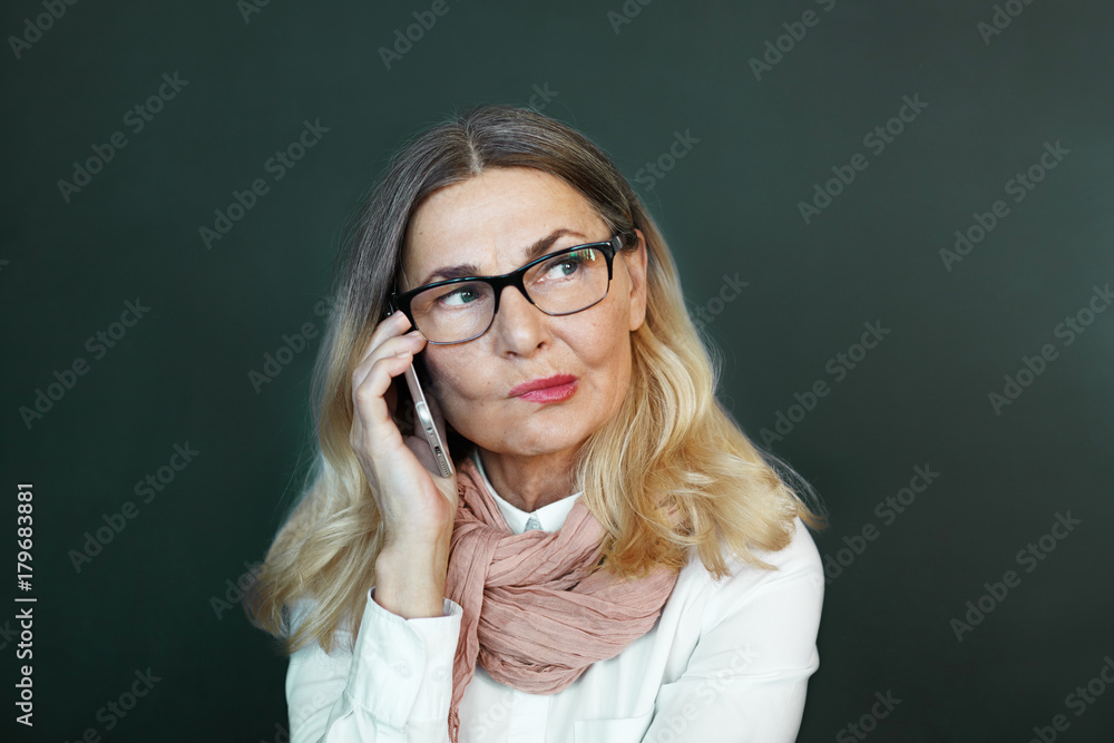Portrait of attractive successful mature businesswoman wearing stylish accessories biting her lips in doubt and indecisiveness, feeling suspicious whilehaving phone conversation at blank office wall Portrait of attractive successful mature businesswoman wearing stylish accessories biting her lips in doubt and indecisiveness, feeling suspicious whilehaving phone conversation at blank office wall