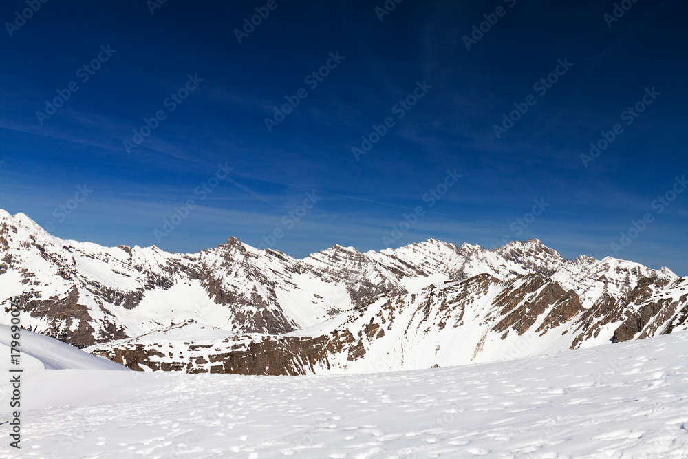 Panorama of mountains ridge in snow forming valley under bright blue sky in sunlight.