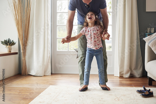 Girl balancing on father’s feet at home, looking up