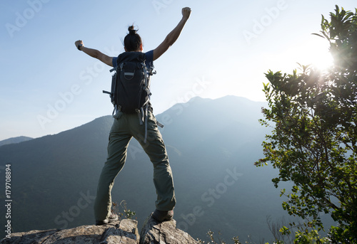 young woman backpacker standing on cliff's edge with raised hands