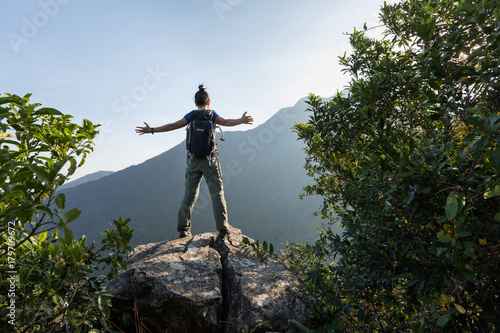young woman backpacker standing on cliff's edge with raised hands