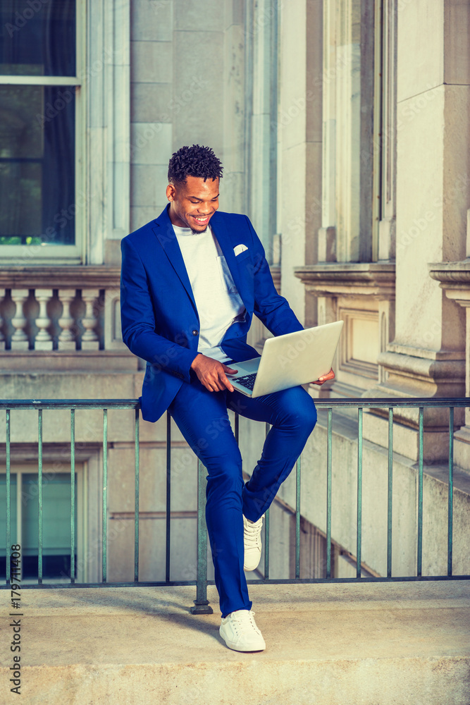 African American college student studying in New wearing blue suit, white sneakers, sitting on railing in vintage office on campus, looking down, working on computer, smiling.. Stock Photo
