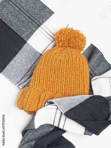 Mustard knit beanie hat with big pom pom and black and white checked scarf isolated on white background. Copy space. Flat lay. Top view