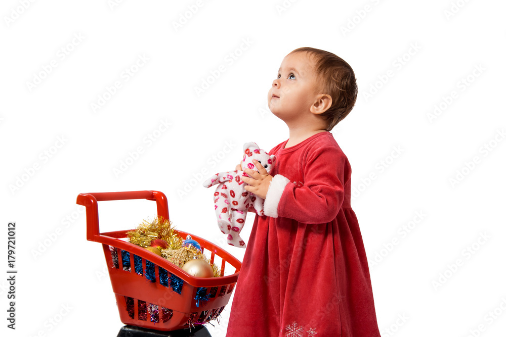 baby in red dress standing near supermarket trolley of colorful Christmas stuff and looking up, isolated on white background. baby in red dress standing near supermarket trolley of colorful Christmas stuff and looking up, isolated on white background.