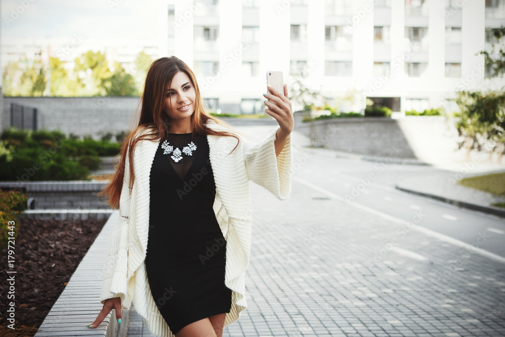 Fototapeta premium Beautiful young fashionable woman in a black short dress and white cardigan posing in the city. Fashion photo