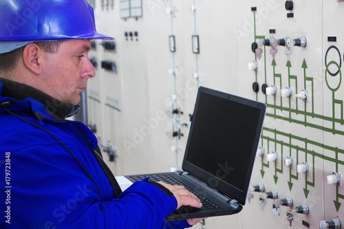 Technician in blue with laptop reading instruments in power plant control center
