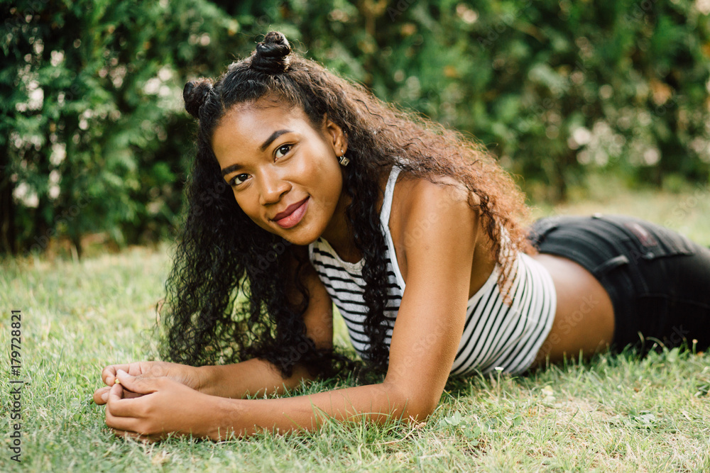 Portrait of a happy young ecuadorian woman with fancy hairstyle like a cat  in striped T- shirt smiling and lying on the grass at park in sunset. Stock  Photo | Adobe Stock