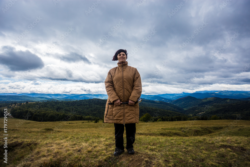 Happy senior tourist woman travel in mountain forest in autumn with dramatic sky on background