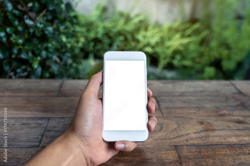 Mockup image of a man's hand holding and using white smart phone with blank screen on wooden table and green nature background Mockup image of a man's hand holding and using white smart phone with blank screen on wooden table and green nature background