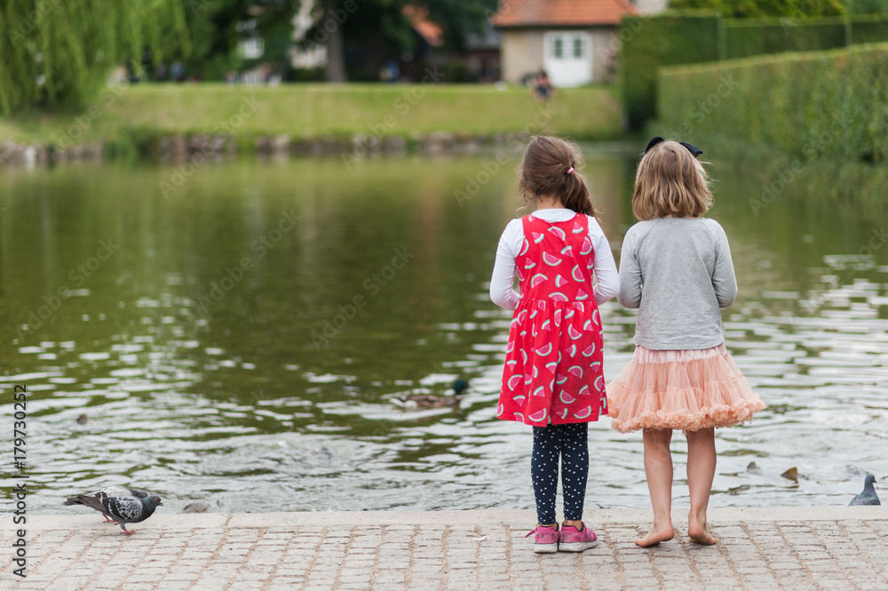 Young girls feeding birds by the city pond. Throwing bread to ducks. Pretty colorful dresses and skirts, long hair. Young girls feeding birds by the city pond. Throwing bread to ducks. Pretty colorful dresses and skirts, long hair.