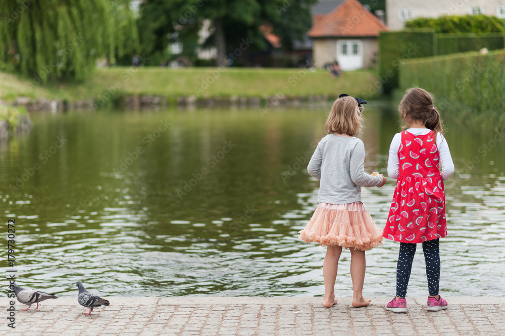 Young girls feeding birds by the city pond. Throwing bread to ducks. Pretty colorful dresses and skirts, long hair. Young girls feeding birds by the city pond. Throwing bread to ducks. Pretty colorful dresses and skirts, long hair.