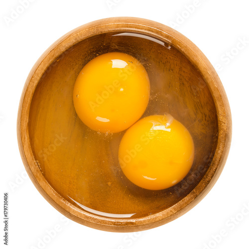 Two raw chicken eggs cracked into a wooden bowl. Yolk and white without eggshells. Common food and versatile ingredient in cooking. Isolated macro food photo close up from above on white background.
