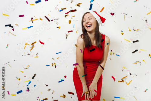 Beautiful caucasian young happy woman wearing red dress and Christmas hat laughing with her eyes closed with pleasure on white background with confetti. Santa girl. New Year holiday 2018 concept