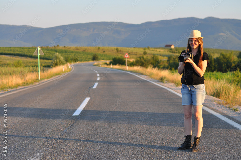 Obraz premium Woman walking with a camera on the middle of the road in mountains. Forty Years old (40 yo) woman on the road