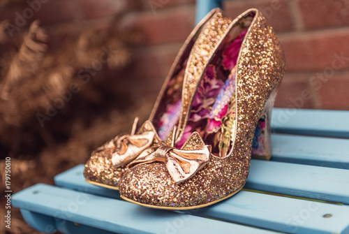 A brides beautiful, golden, glittery, unique, sexy pair of individual stylish wedding shoes, on a blue wooden surface outdoors.