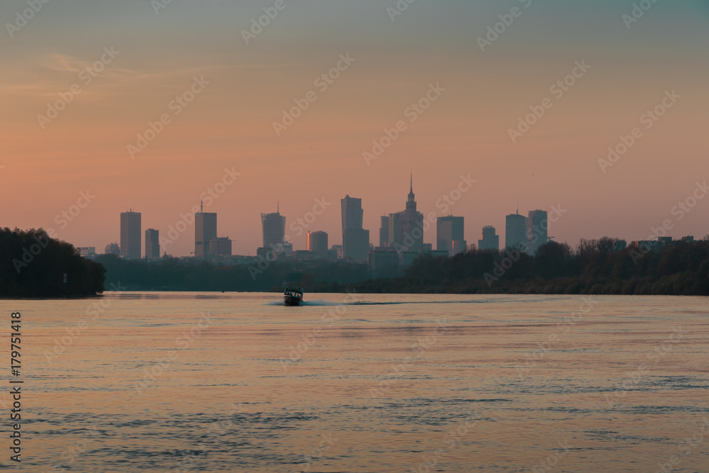 Warsaw skyline with skyscrapers and a boat sailing on the Vistula River in red orange light of the setting sun at dusk
