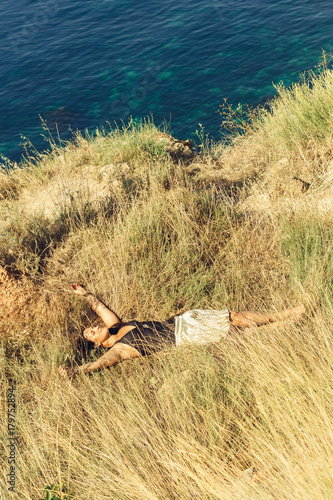 A beautiful young woman lies in a field on a cliff near a clear blue sea and enjoys the sound of waves on a summer vacation by the ocean. Harmony and silence. Meditation.