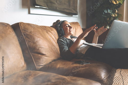 Fotografie Attractive young happy girl listening to music in headphones with pleasure at coworking office, lying on sofa, using laptop and resting